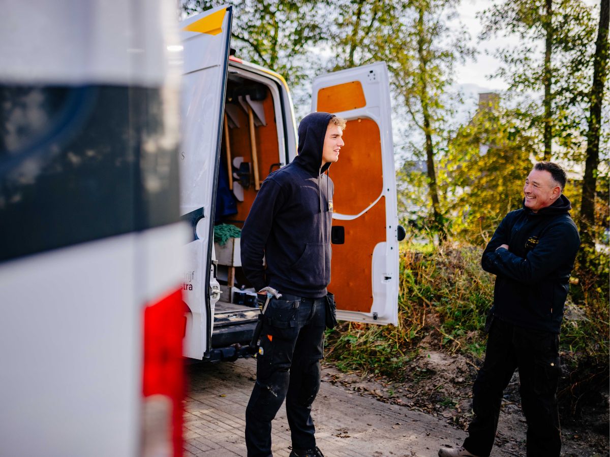 Twee collega’s van U. Veenstra staan naast de open werkbus en overleggen ontspannen op de bouwlocatie, met gereedschap en natuur op de achtergrond.