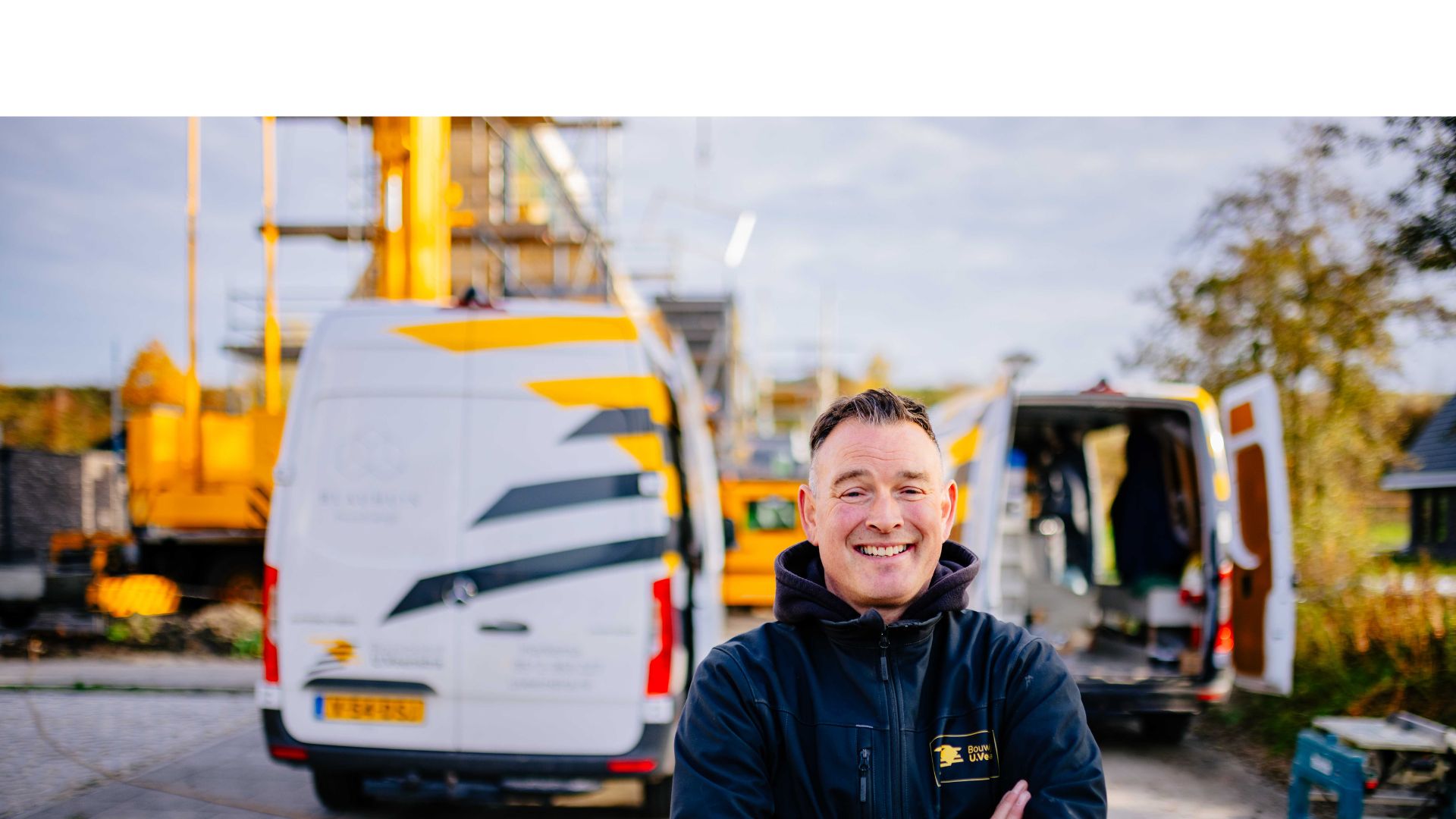 Medewerker van Bouwbedrijf U. Veenstra poseert trots op de bouwplaats voor het herkenbare wagenpark en lopende werkzaamheden