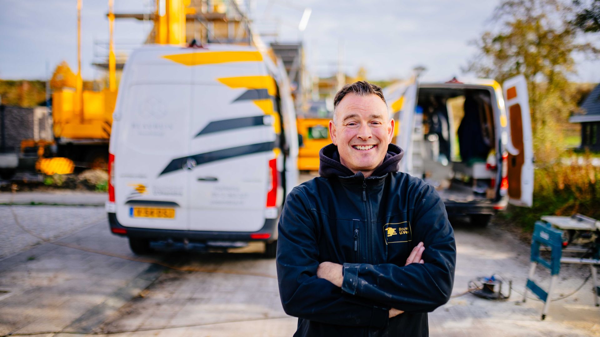 Monteur van Bouwbedrijf U. Veenstra staat met gevouwen armen op de bouwplaats, met bedrijfsbussen en materieel op de achtergrond