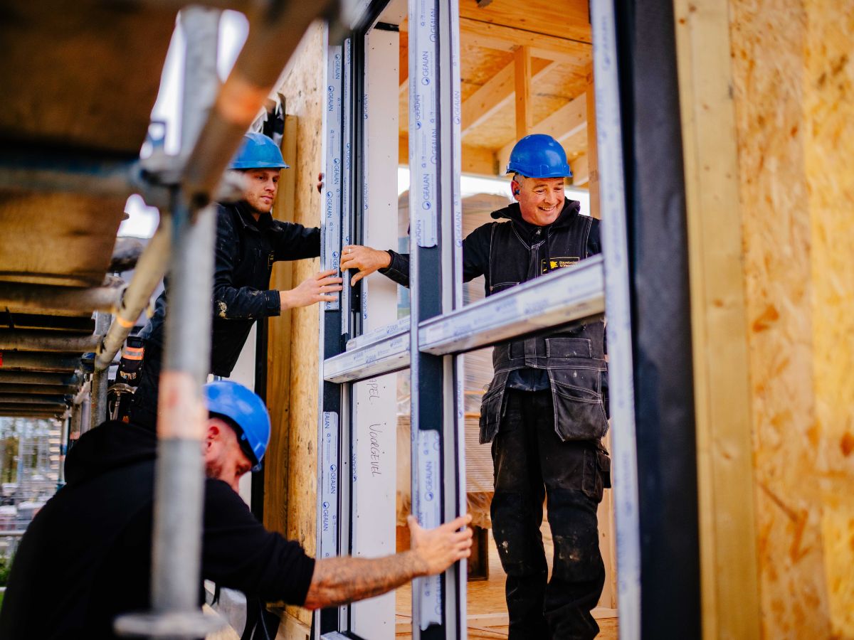 Collega’s van U. Veenstra plaatsen samen raamkozijnen in een houtskeletbouwwoning, terwijl ze nauwkeurig afstemmen en samenwerken op de bouwplaats.