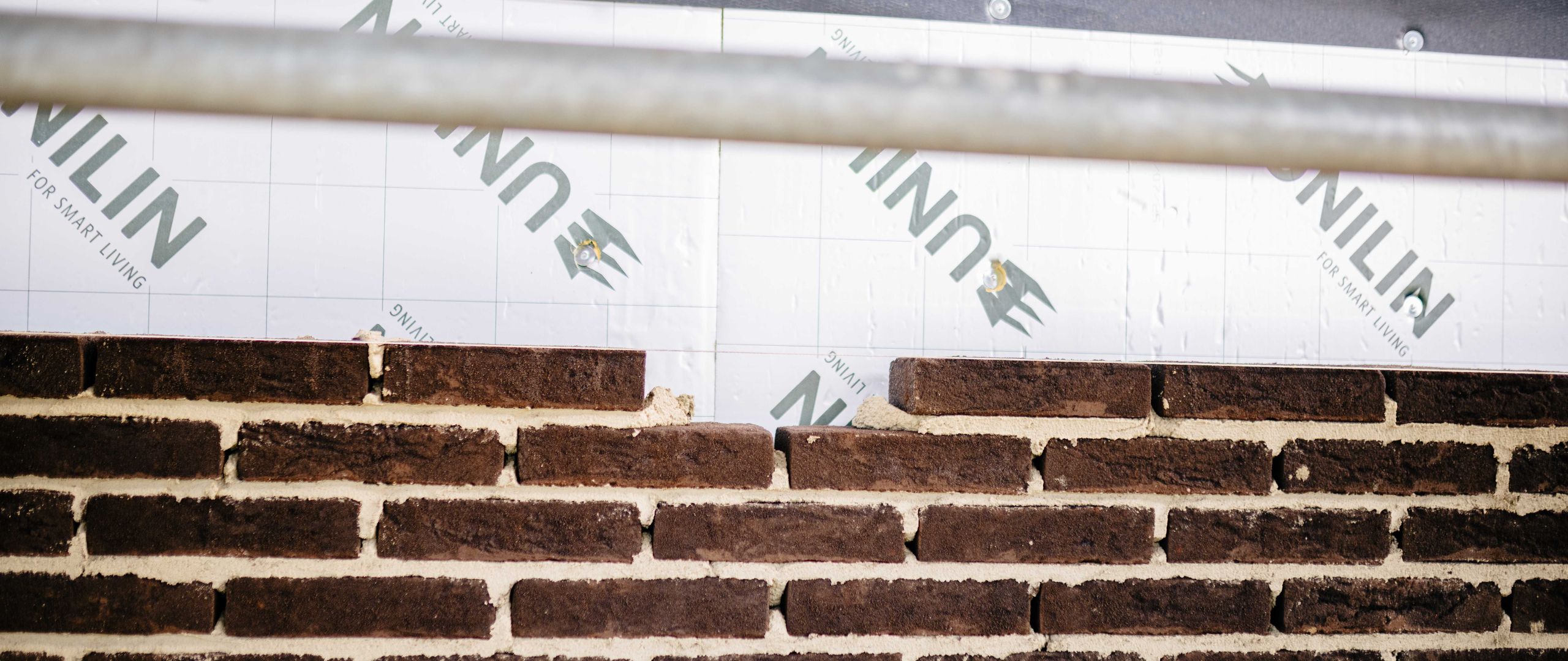 Close-up van een half opgemetselde bakstenen muur, waarbij de bovenste laag nog wordt opgebouwd tegen isolatieplaten tijdens de ruwbouwfase van een woning.