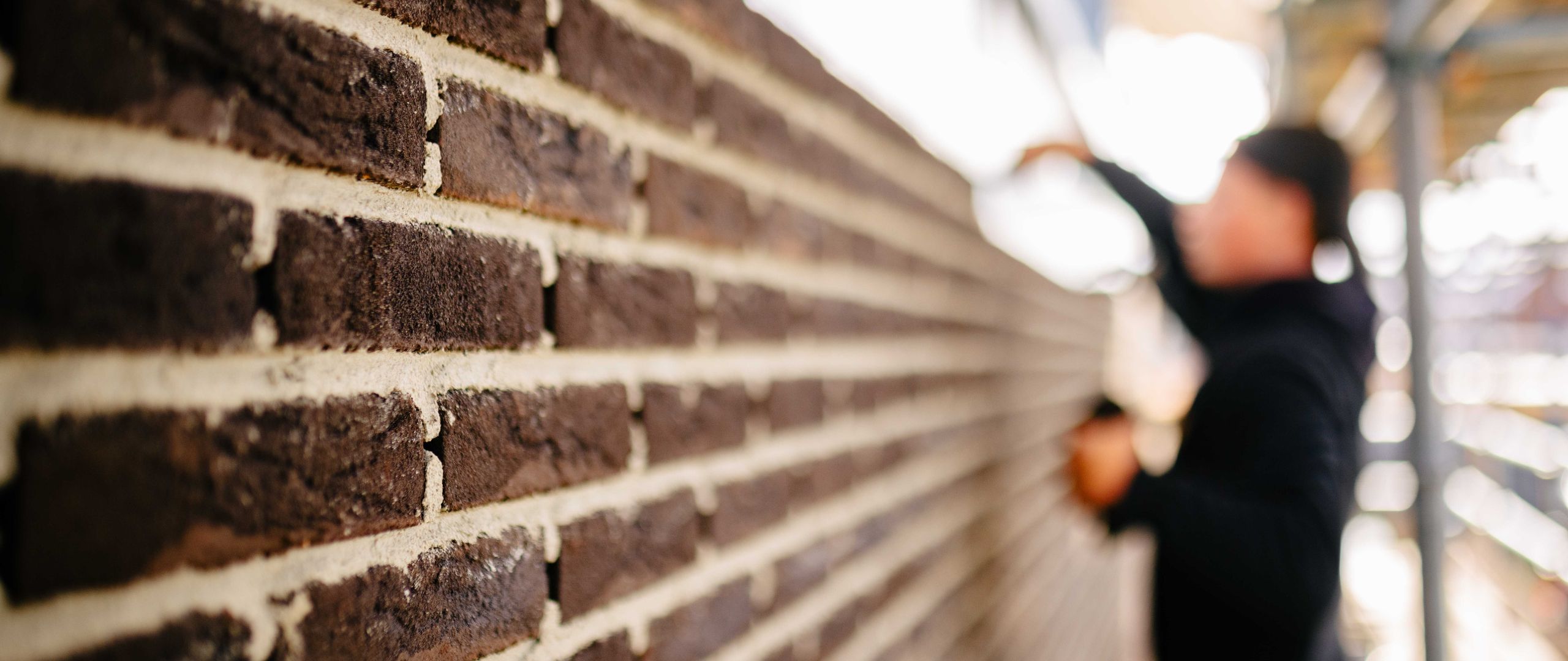 Close-up van strak gemetselde bakstenen met lichte voegen, terwijl op de achtergrond een vakman op de steiger aan het werk is, onscherp in beeld.