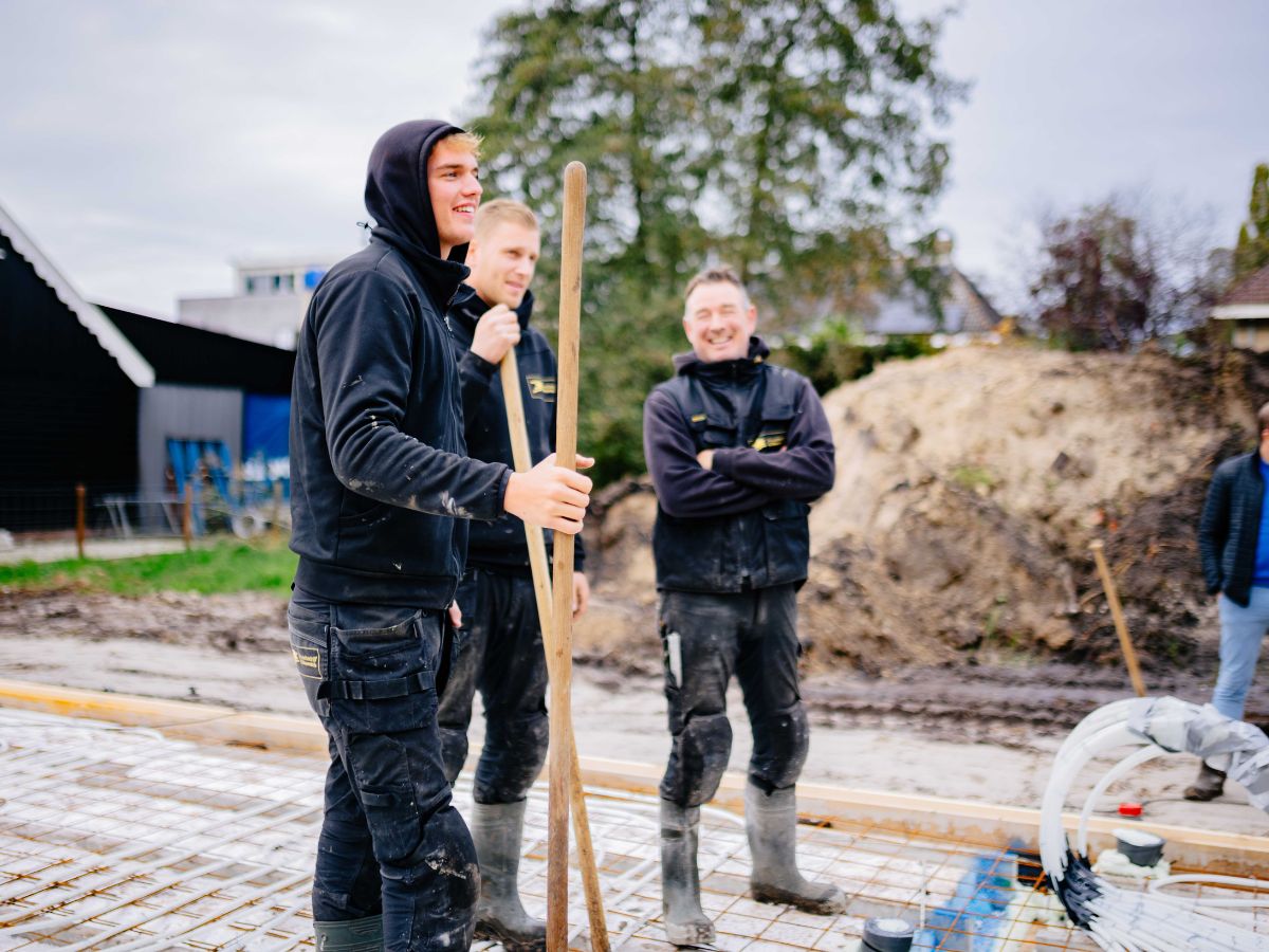 Drie collega’s van U. Veenstra werken samen op de bouwplaats; een jonge medewerker staat voorop met een hark, terwijl collega’s op de achtergrond toekijken bij het voorbereiden van de fundering.