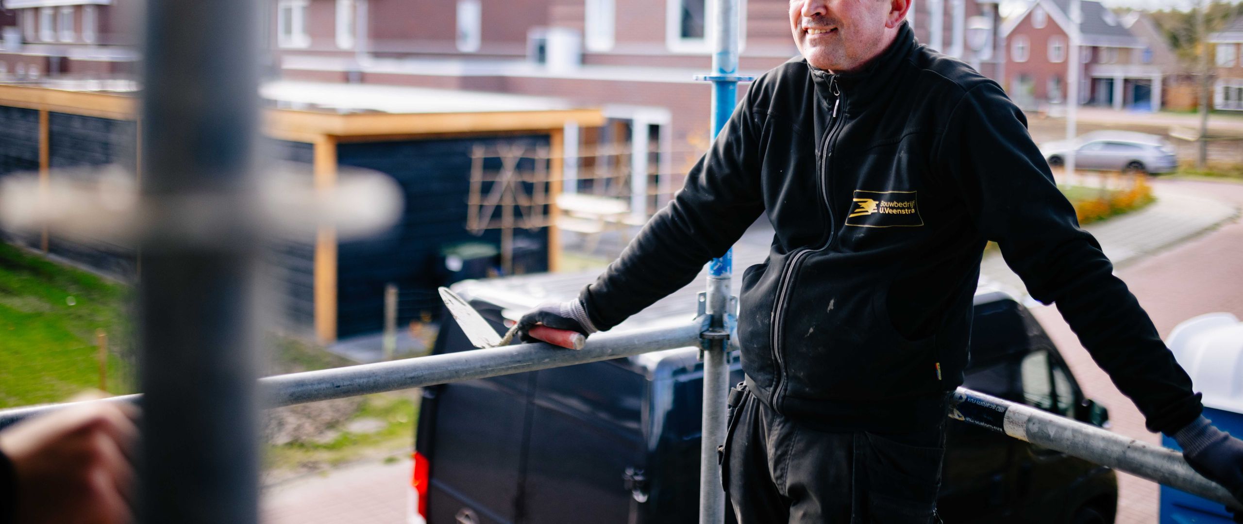 Een medewerker van U. Veenstra staat ontspannen op een steiger en leunt op de reling, met een bedrijfsbus en nieuwbouwwoningen zichtbaar op de achtergrond van de bouwplaats.