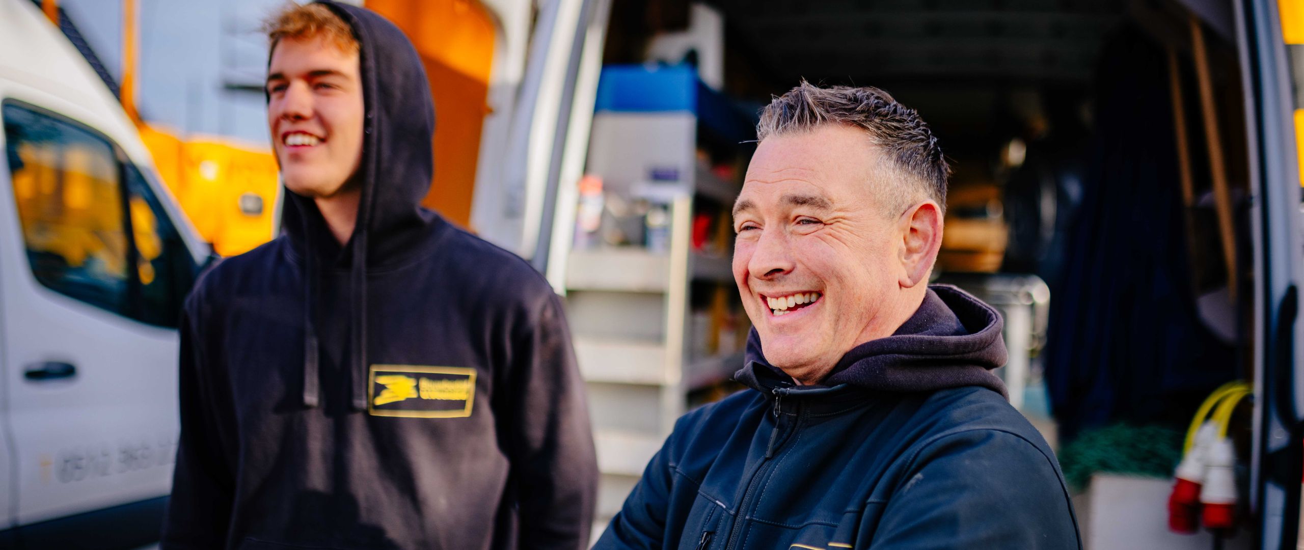 Twee collega’s van Bouwbedrijf U. Veenstra staan lachend bij een servicebus op de bouwplaats, zichtbaar in gesprek en ontspannen.