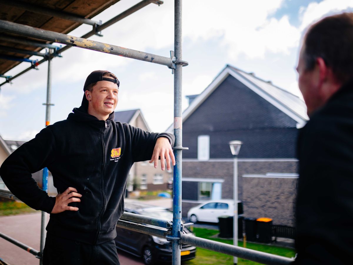 Een medewerker van U. Veenstra leunt ontspannen tegen een steiger en praat met een collega, met op de achtergrond moderne nieuwbouwwoningen in een woonwijk.